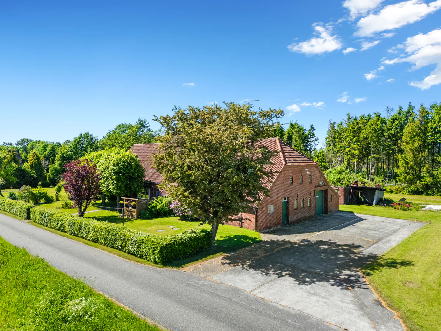 A red brick farmhouse with a tiled roof, surrounded by lush green trees, a manicured hedge, and a paved driveway.