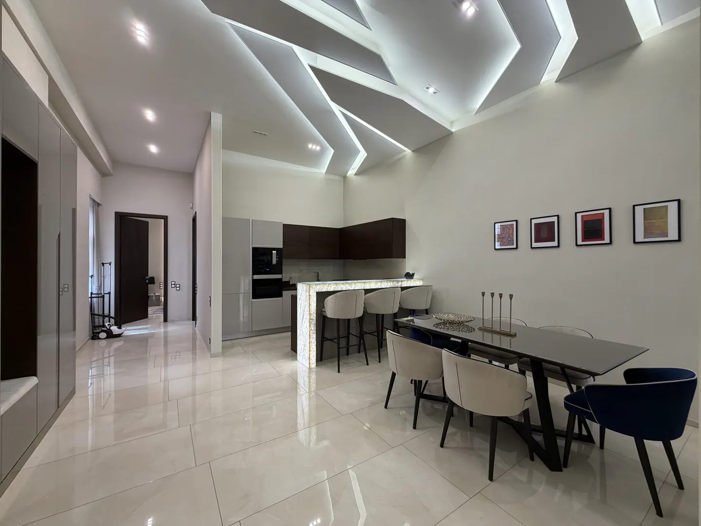 A modern, open-concept kitchen and dining area with white tile floors and geometric ceiling lighting. A black dining table with beige and blue chairs sits near a kitchen island with bar stools.