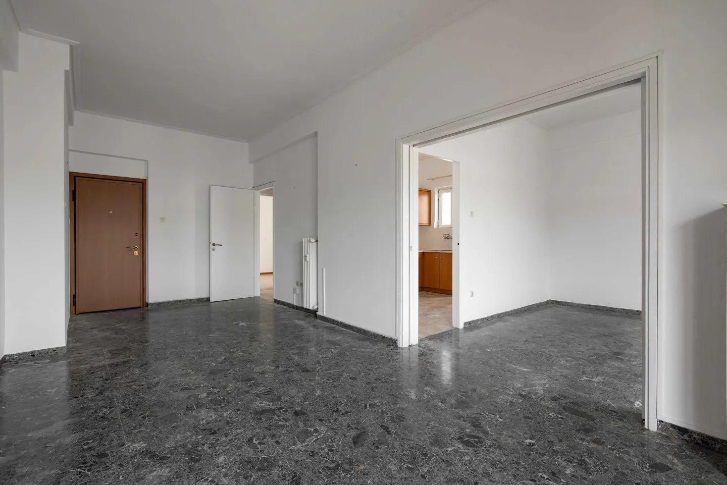Empty room with gray marble floor, white walls, and brown front door. Kitchen visible through doorway.