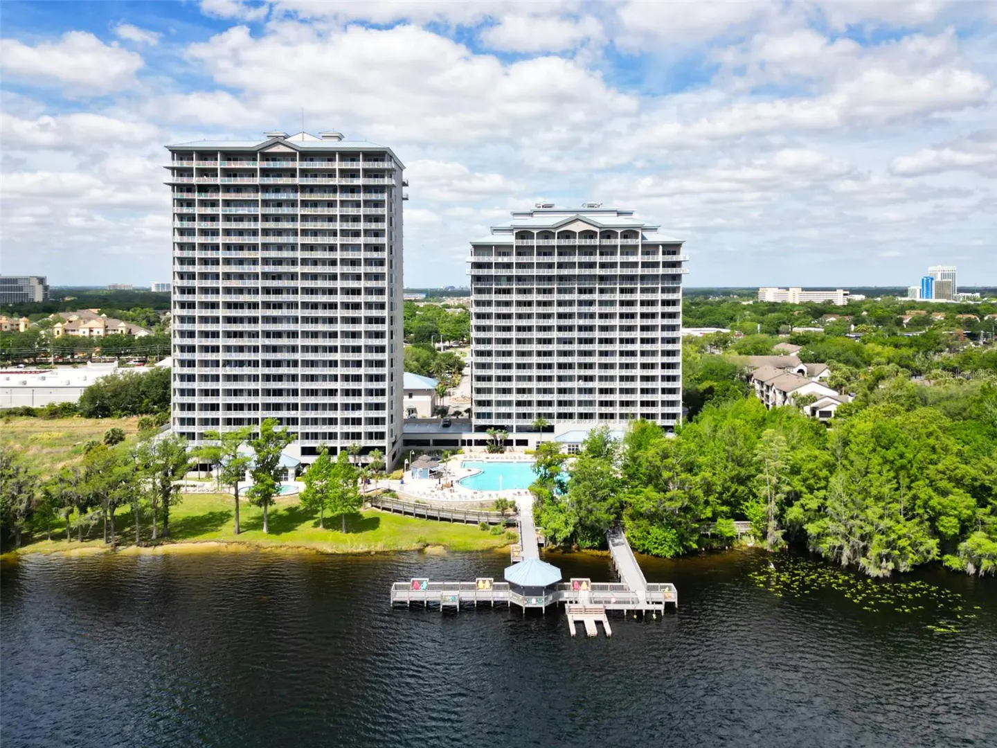 Aerial view of two tall, white condo buildings on a lake, with a wooden dock and gazebo in the water. A blue pool is visible between the buildings.