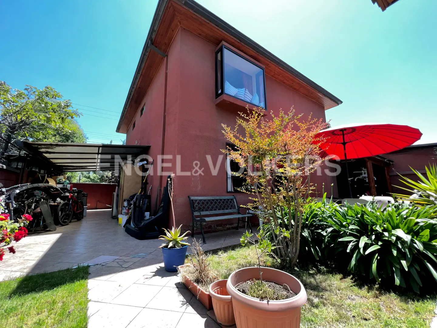 Exterior view of a two-story red house with a garden, bench, and red umbrella on a sunny day.