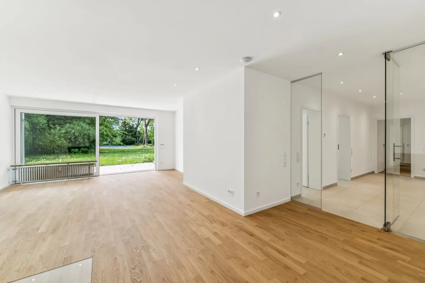 Bright, empty room with hardwood floors, white walls, and a large sliding glass door to a green yard. Glass doors lead to other rooms.