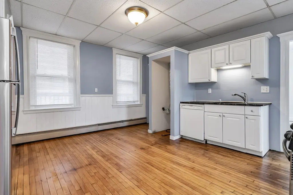 A bright kitchen with hardwood floors, white cabinets, and stainless steel appliances. Two windows with blinds let in natural light.