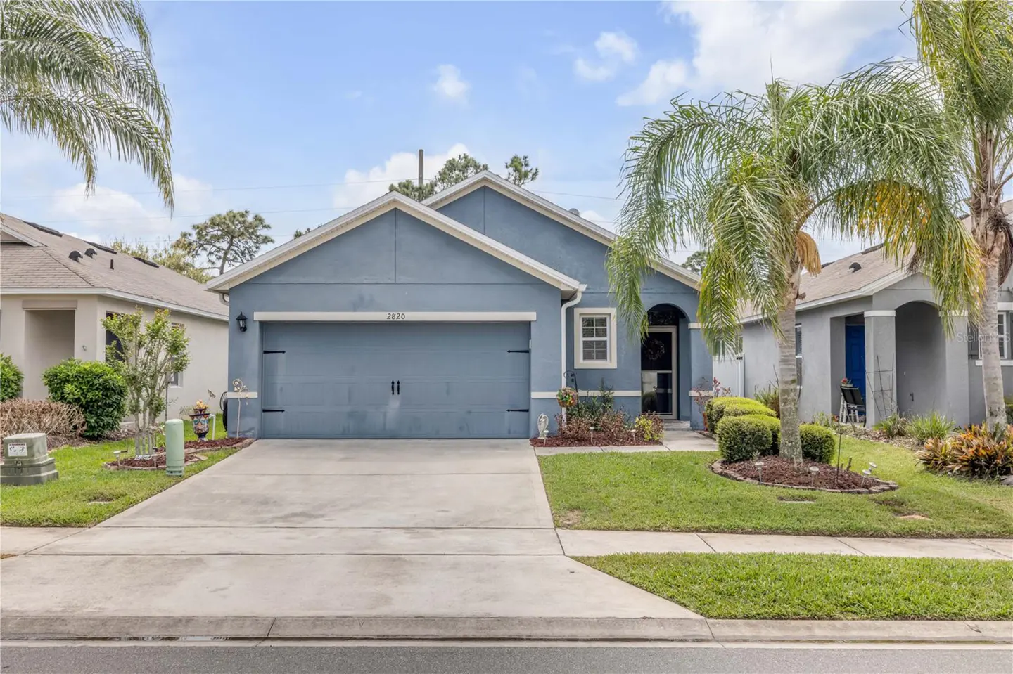 A blue single-story house with a garage, a concrete driveway, and palm trees in the front yard.