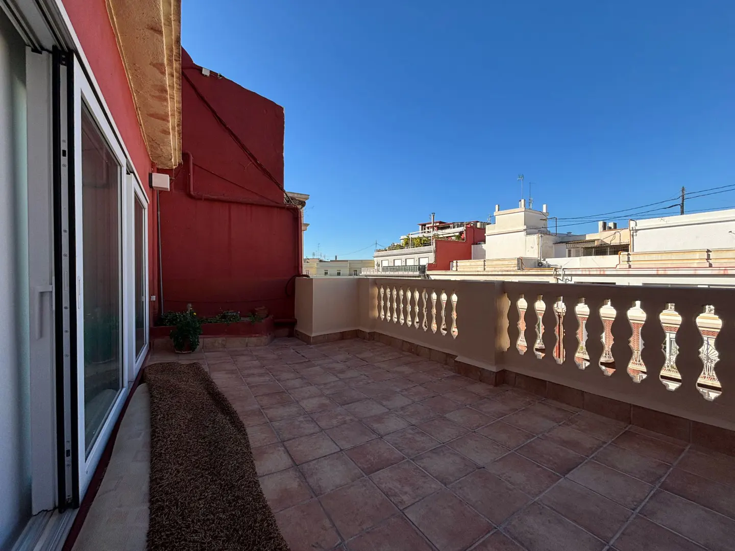 A rooftop terrace with terracotta tiles, a decorative white railing, and a red building in the background under a clear blue sky.