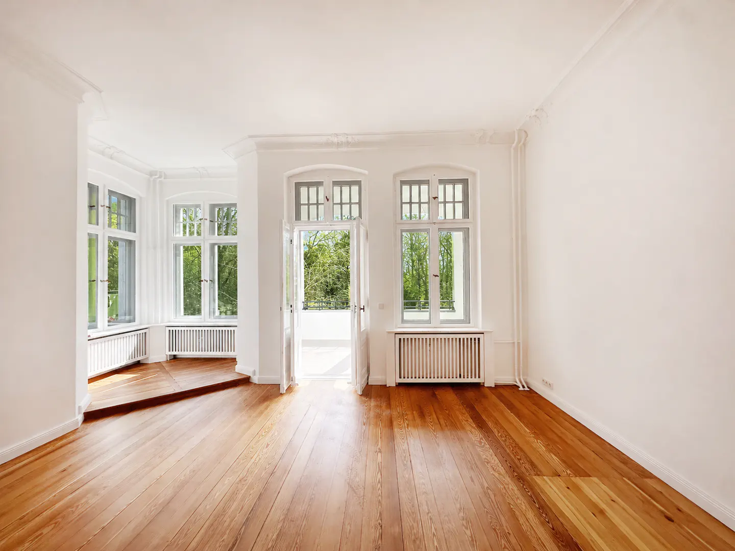 Bright, empty room with hardwood floors, white walls, and large windows overlooking greenery. Open doors lead to a balcony.