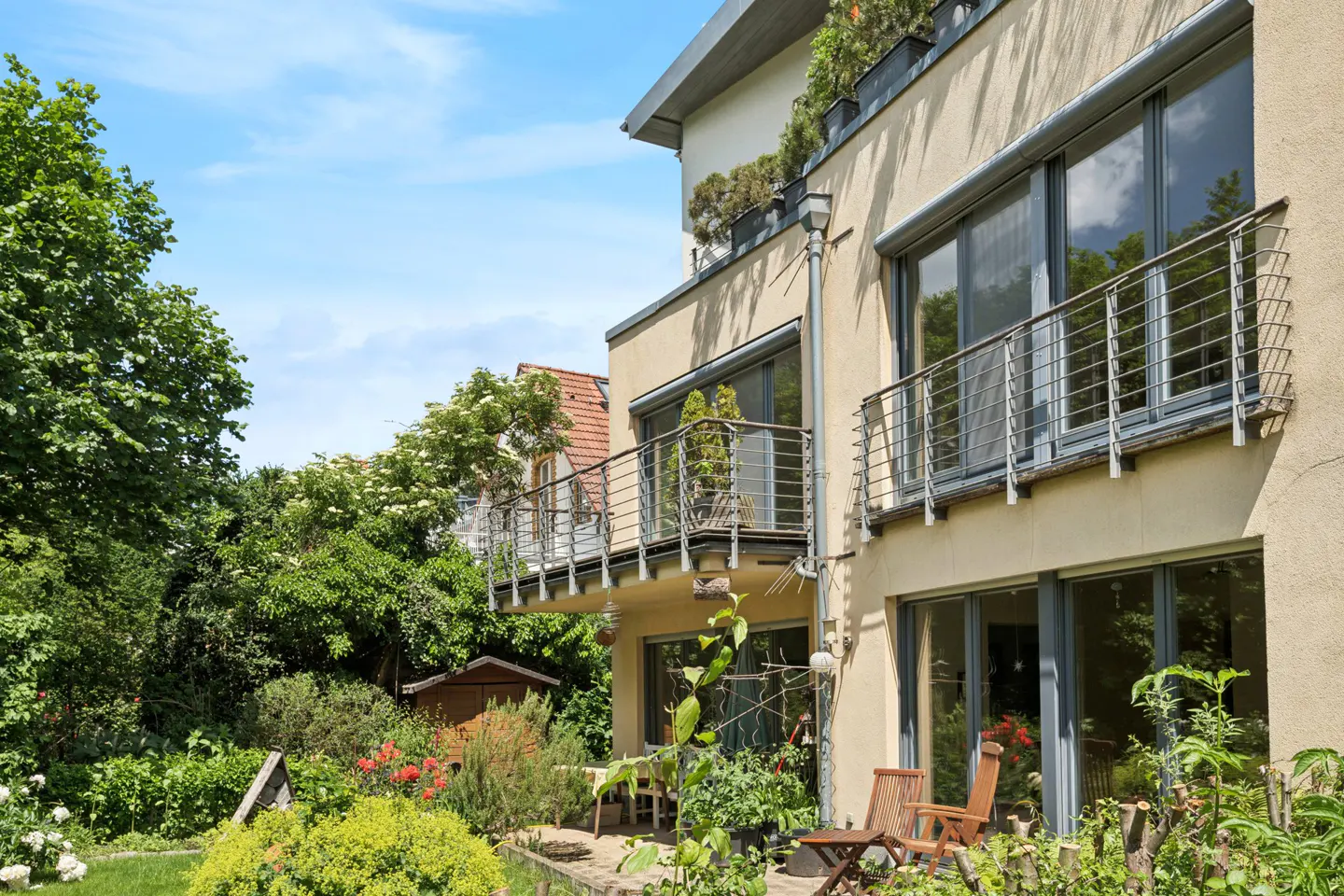 Modern beige house with balconies, large windows, and a lush green garden under a bright blue sky.