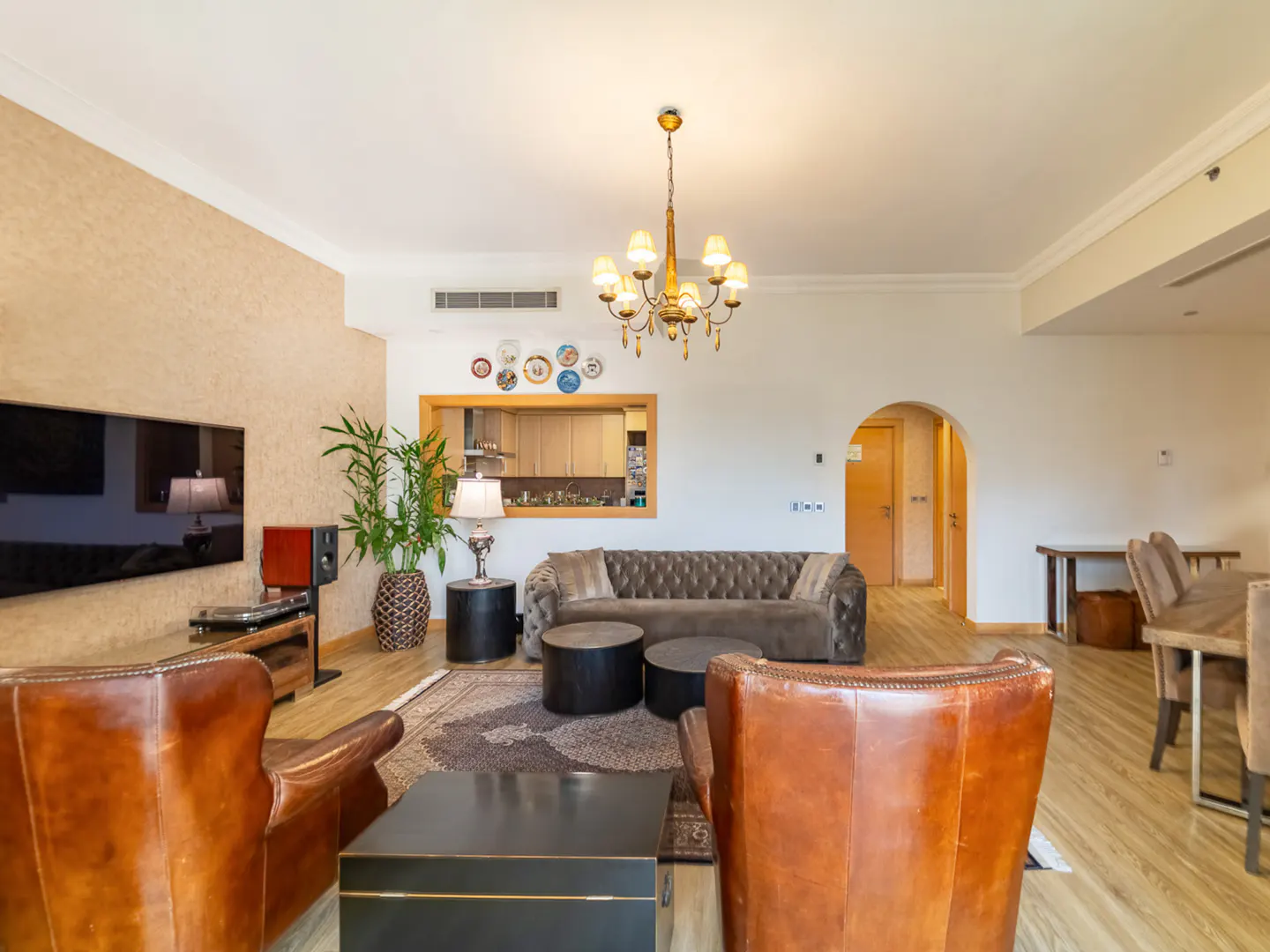 Living room with brown leather armchairs, a tufted sofa, a chandelier, and a view into the kitchen through a wall cutout.