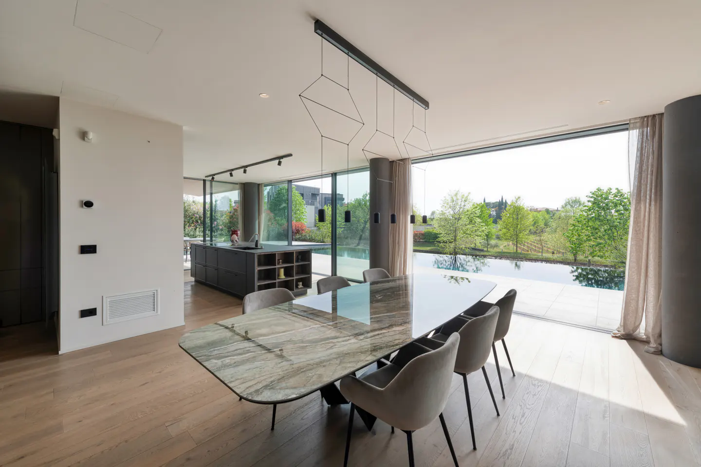 Bright, modern dining room with a long marble table, gray chairs, and geometric light fixture. Large windows overlook a pool and green landscape.