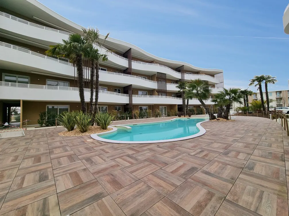 Modern apartment building with a pool. Palm trees and plants surround the pool area. The building has white balconies and brown accents.