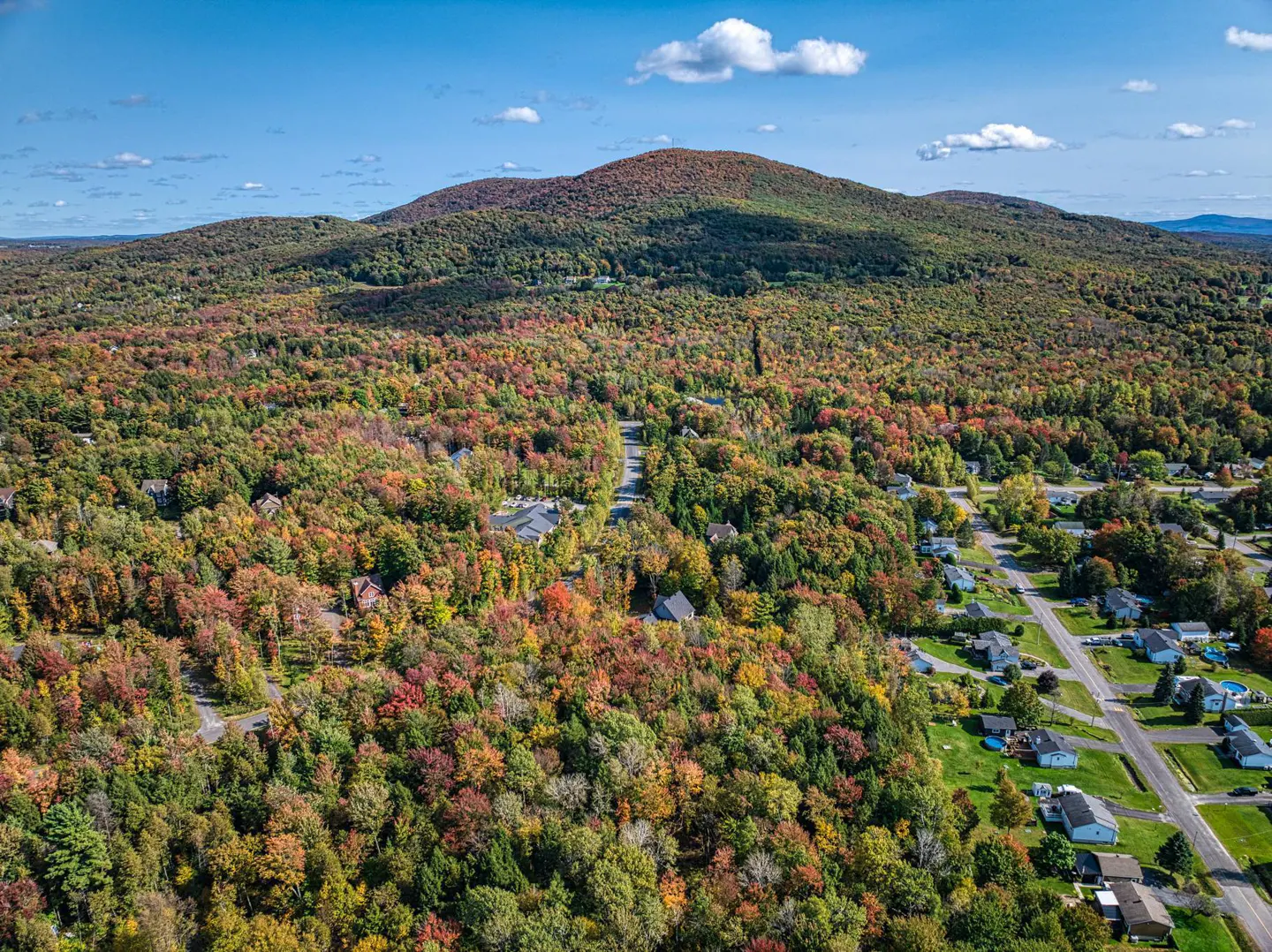 Aerial view of a mountain and neighborhood with fall foliage. Trees are red, orange, and green. Blue sky with clouds.