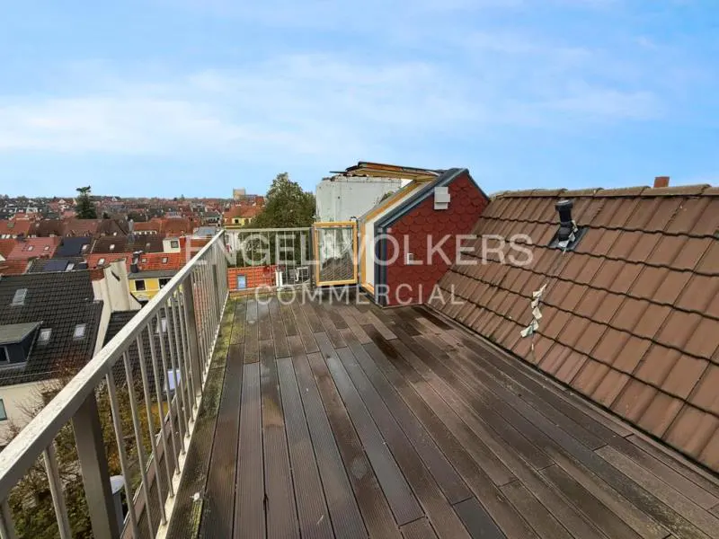 Rooftop view with wood deck, metal railing, and red-tiled roofs. Cityscape visible in the background under a blue sky.