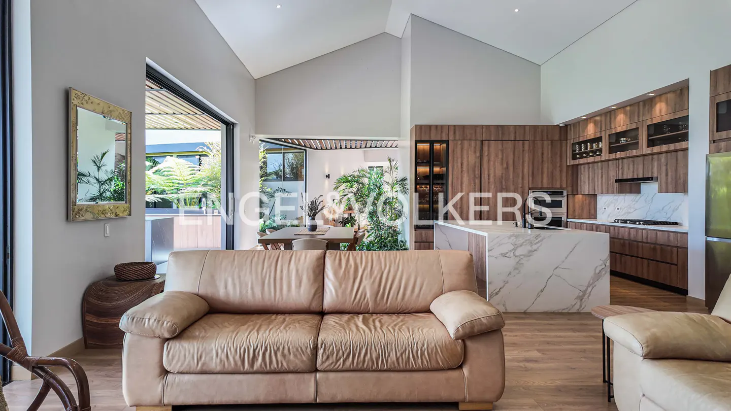 Open-concept living space with a tan leather sofa, wood cabinets, and marble island. Outdoor dining visible through sliding glass doors.