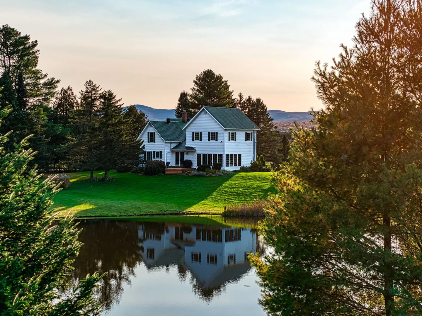 White farmhouse with a green roof, surrounded by trees and a pond reflecting the house.