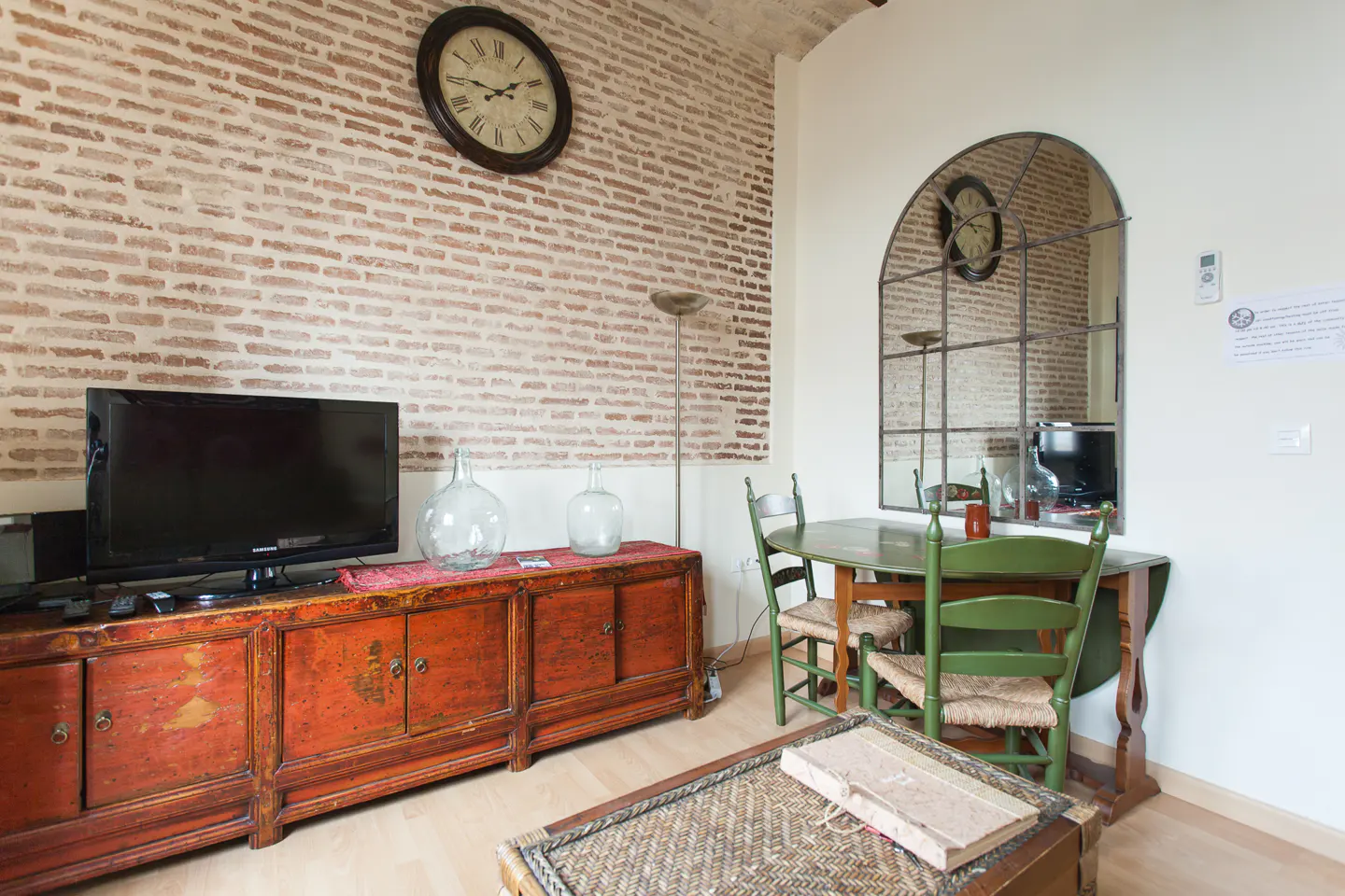 Living room with brick wall, TV on red cabinet, round table with green chairs, and a large arched mirror.