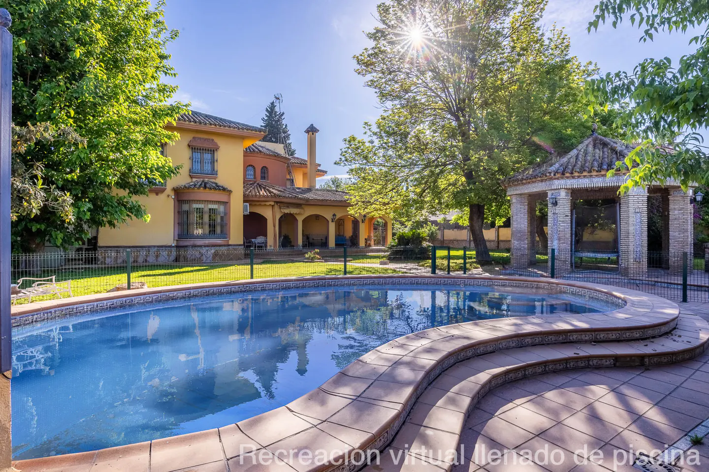 Exterior view of a yellow house with a blue pool, a gazebo, and green trees under a sunny sky.
