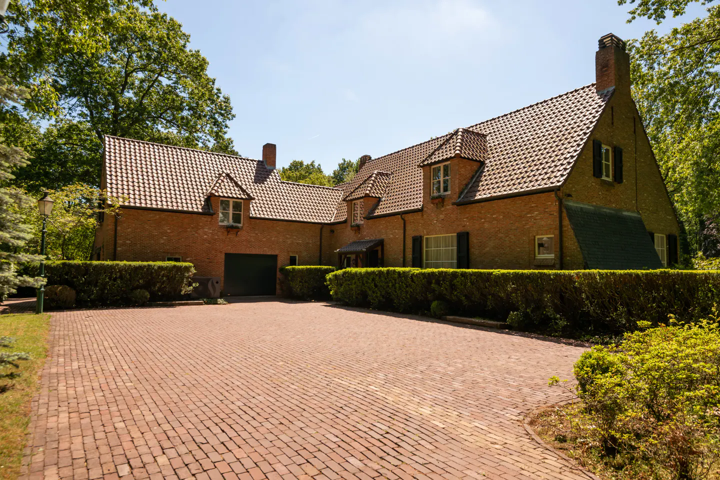 Exterior view of a brick house with a red brick driveway, green bushes, and a tiled roof on a sunny day.