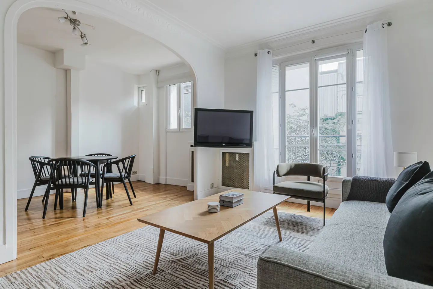 Bright living room with hardwood floors, white walls, and large windows. A gray sofa, wood table, and black dining set furnish the space.