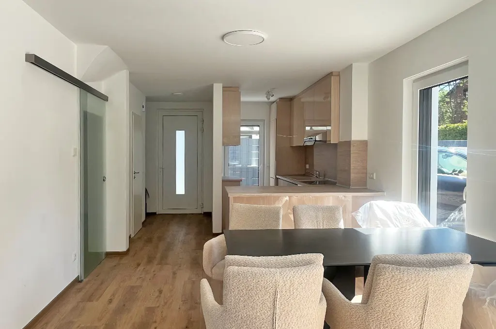 Bright, modern apartment interior with a black dining table, beige chairs, and light wood kitchen cabinets. A sliding glass door is on the left.