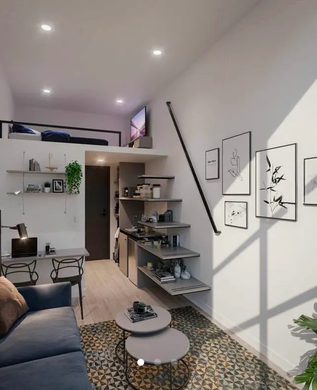 A modern studio apartment with a loft bed, desk, blue sofa, and stairs with storage leading to the loft. Artwork adorns the white walls.