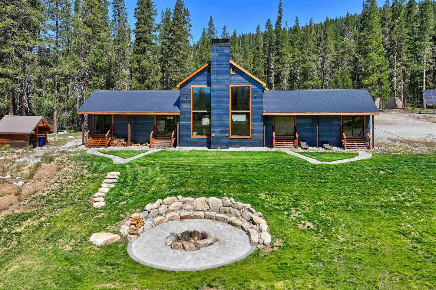 Blue house with a black roof, a stone fire pit, and a green lawn surrounded by tall green trees.
