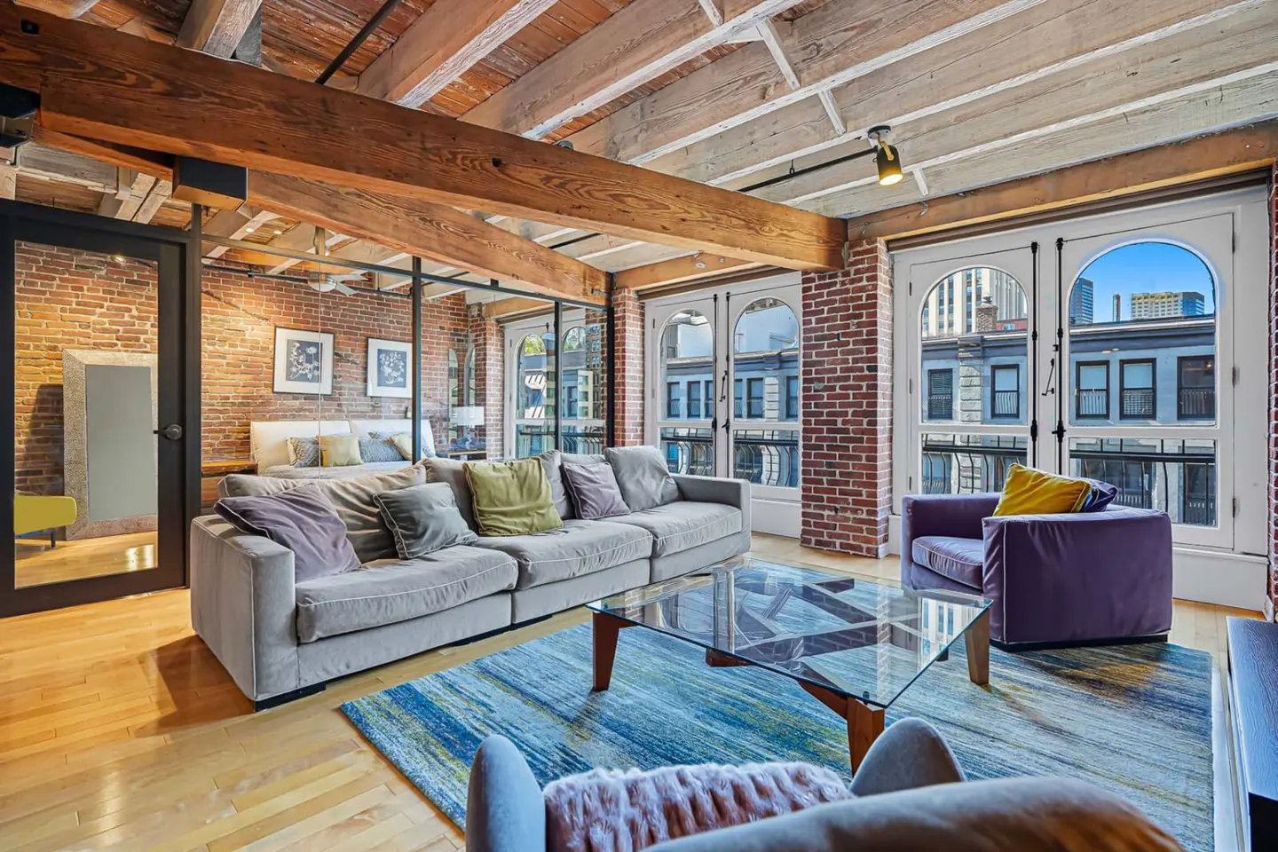 Living room with exposed brick and wood beams. A gray sofa, glass table, and blue rug furnish the space. Large windows offer city views.