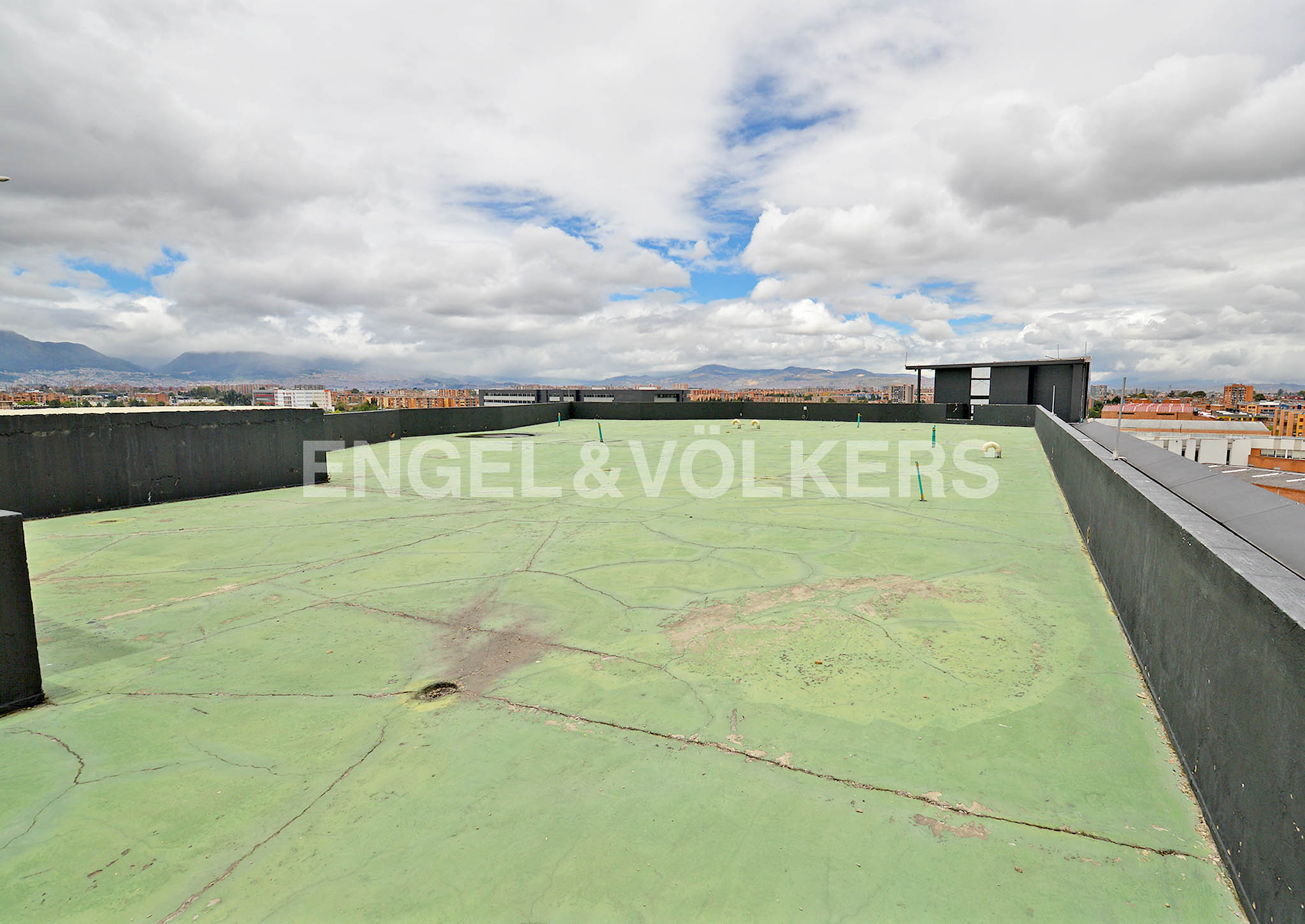 Rooftop view of a green, cracked surface with black walls, set against a cityscape and cloudy sky.