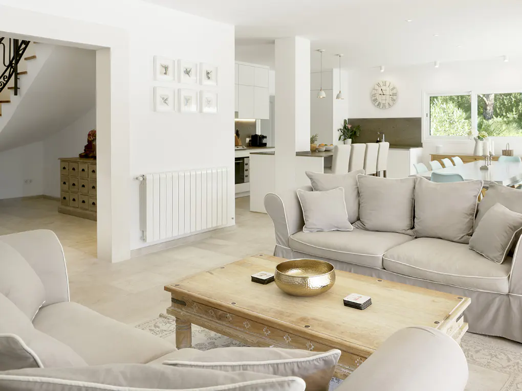Bright, open-concept living room with beige sofas, a wooden coffee table, and a view into the kitchen and dining area. White walls and natural light.