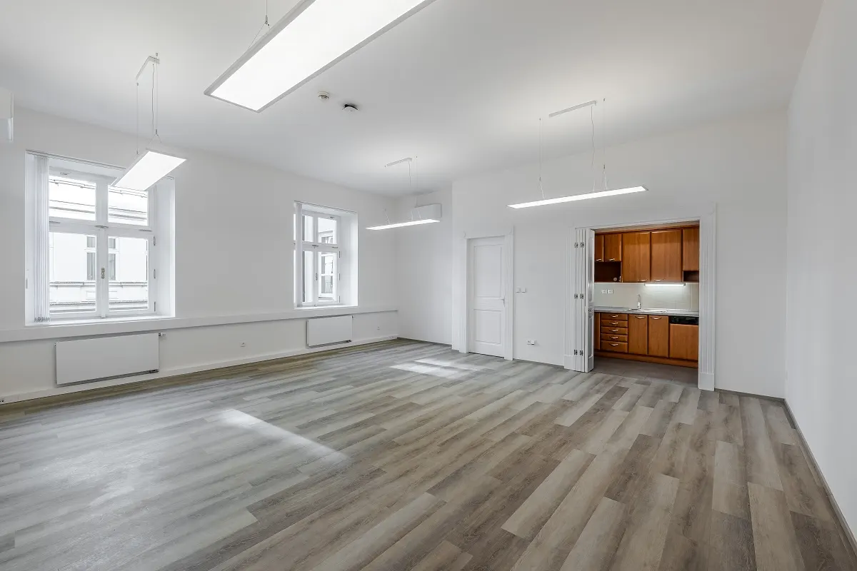 Bright, empty room with gray wood floors, white walls, and two windows. A small kitchen with wood cabinets is visible through an open doorway.