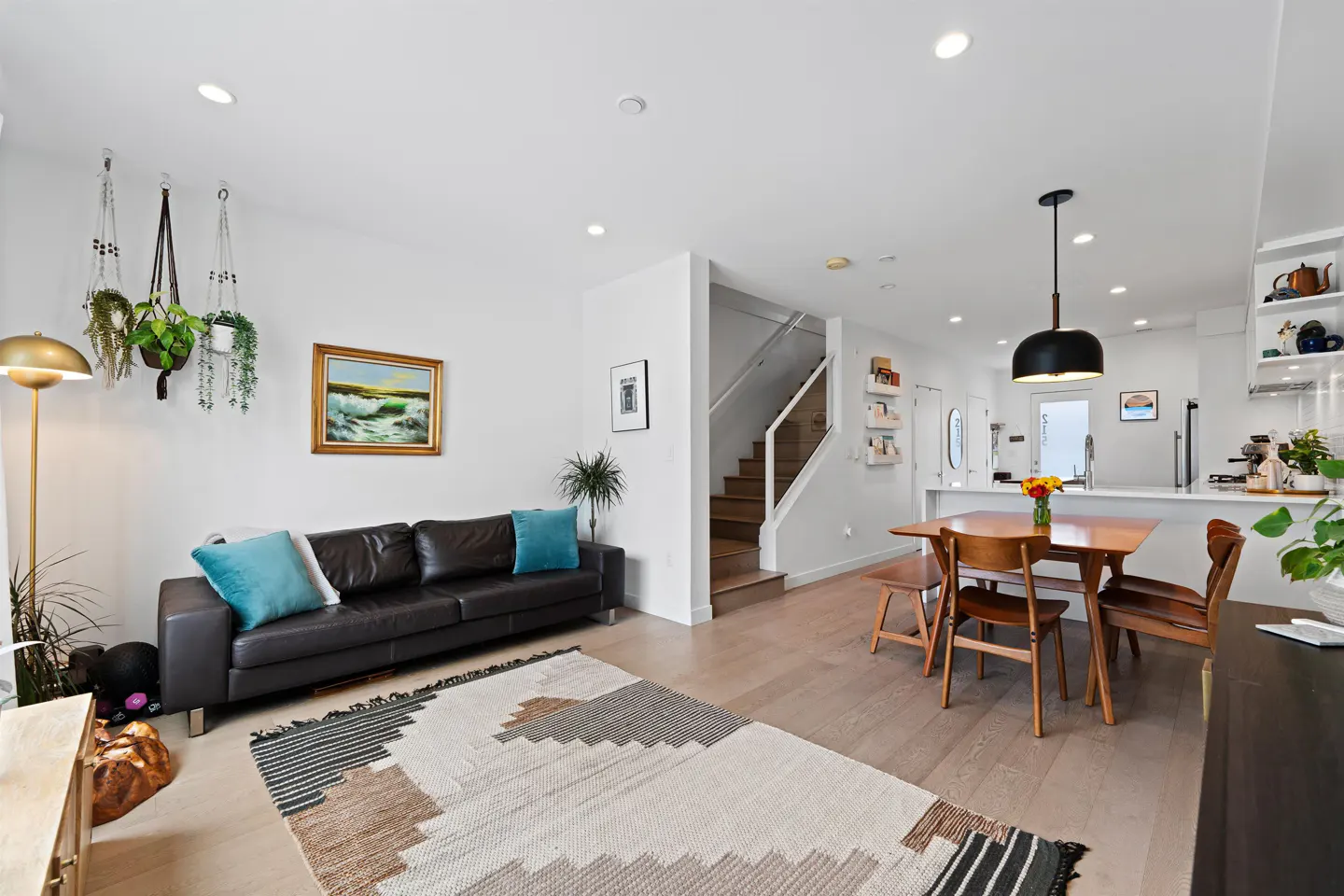 Bright, open-concept living space with a brown leather sofa, dining table, and stairs. Plants and artwork add color.
