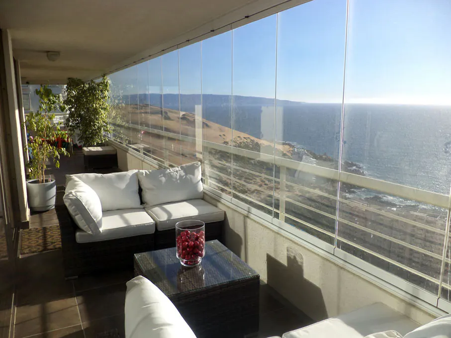 A sunlit balcony with glass walls overlooks the ocean. White sofas and a dark table furnish the space.