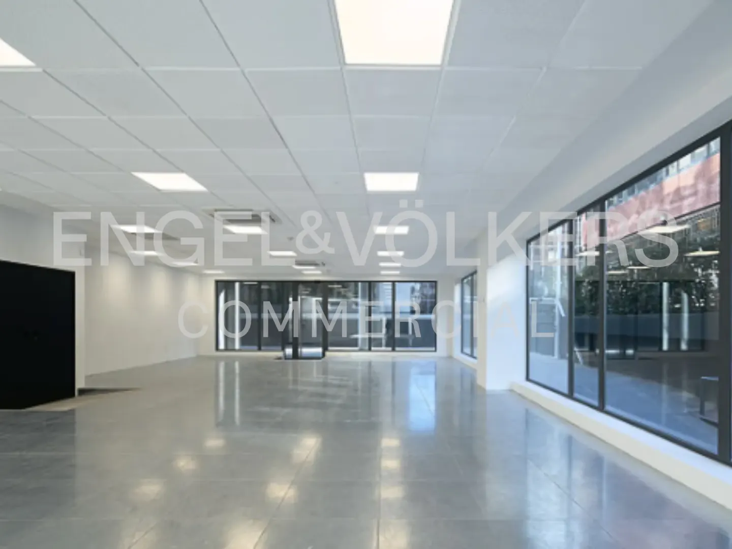 Bright, empty commercial space with white walls, gray floors, and large windows. Square ceiling lights illuminate the room.