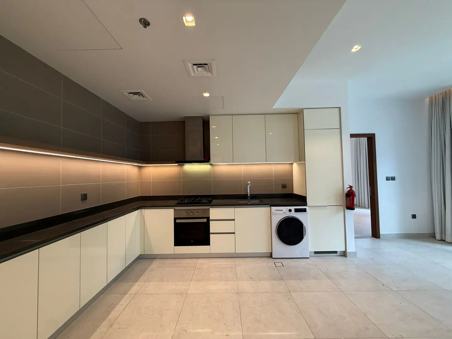 Modern kitchen with cream cabinets, dark countertops, built-in oven, stove, and a white washing machine on tiled floors.