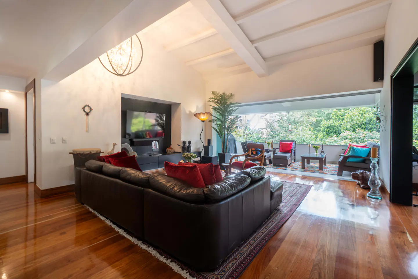 Bright living room with hardwood floors, a brown leather sofa with red pillows, and a balcony with outdoor seating.