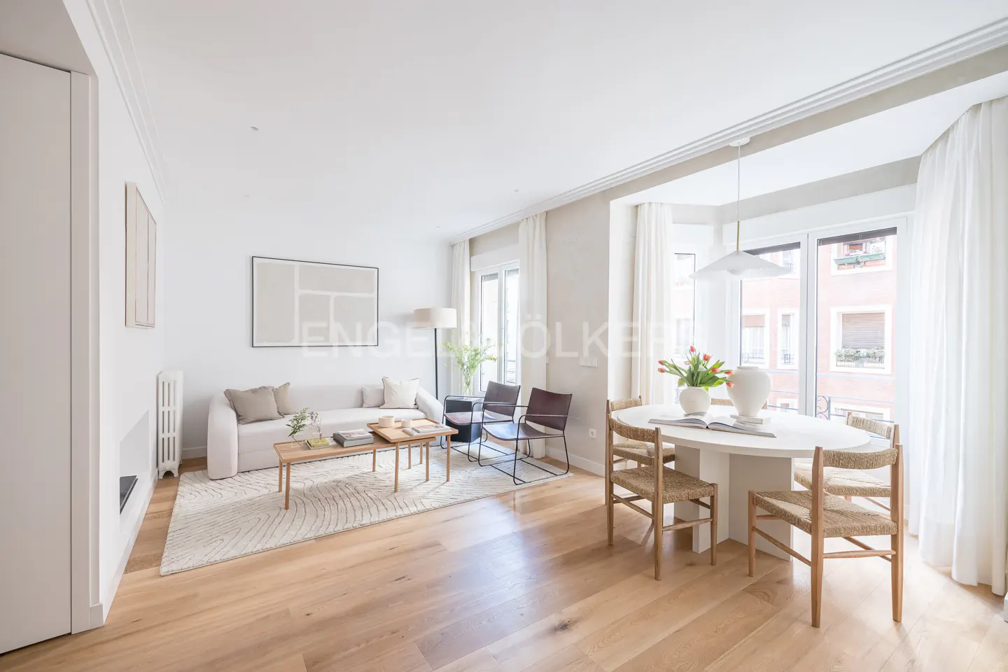 Bright living and dining room with white walls, light wood floors, a beige sofa, and a round table with woven chairs.