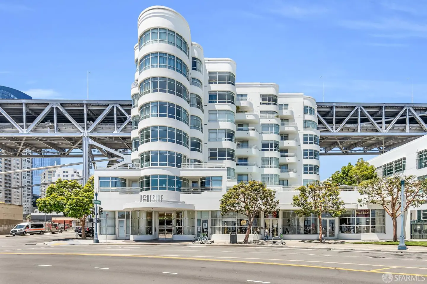 Exterior of a white, art deco apartment building called "Portside" under a bridge on a sunny day.
