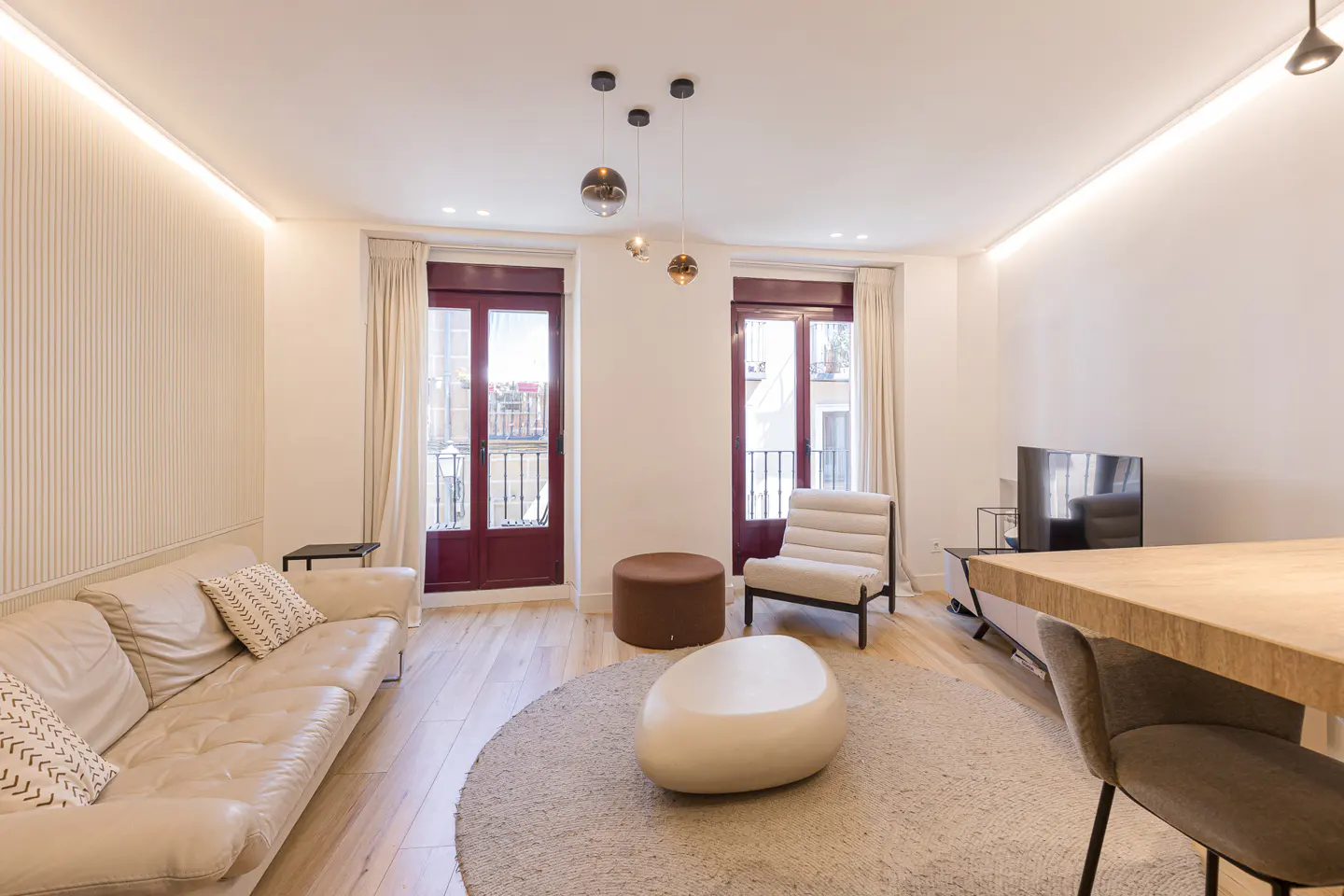 Modern living room with a beige sofa, round rug, pebble-shaped coffee table, and two balcony doors with dark red frames.