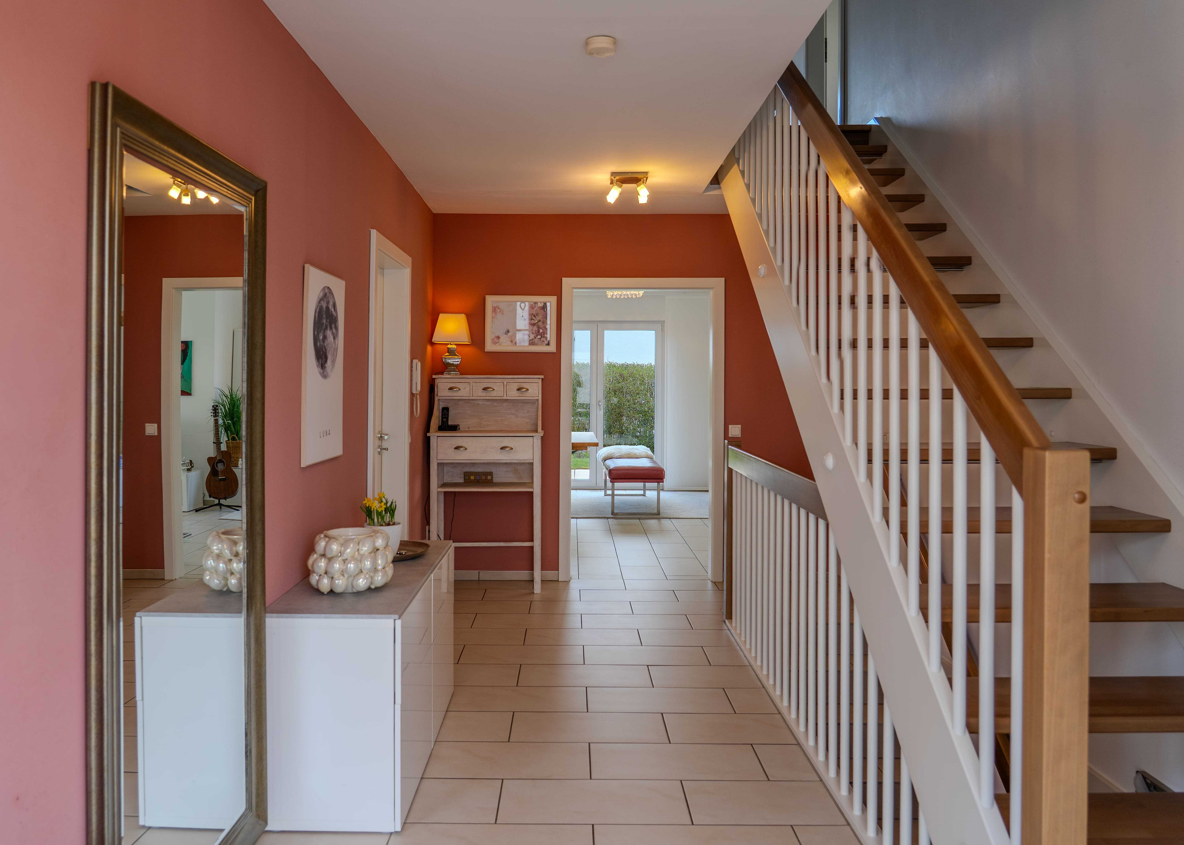 A home's entryway with tile floors, pink and orange walls, and a staircase with white railings. A mirror and white cabinet are on the left.