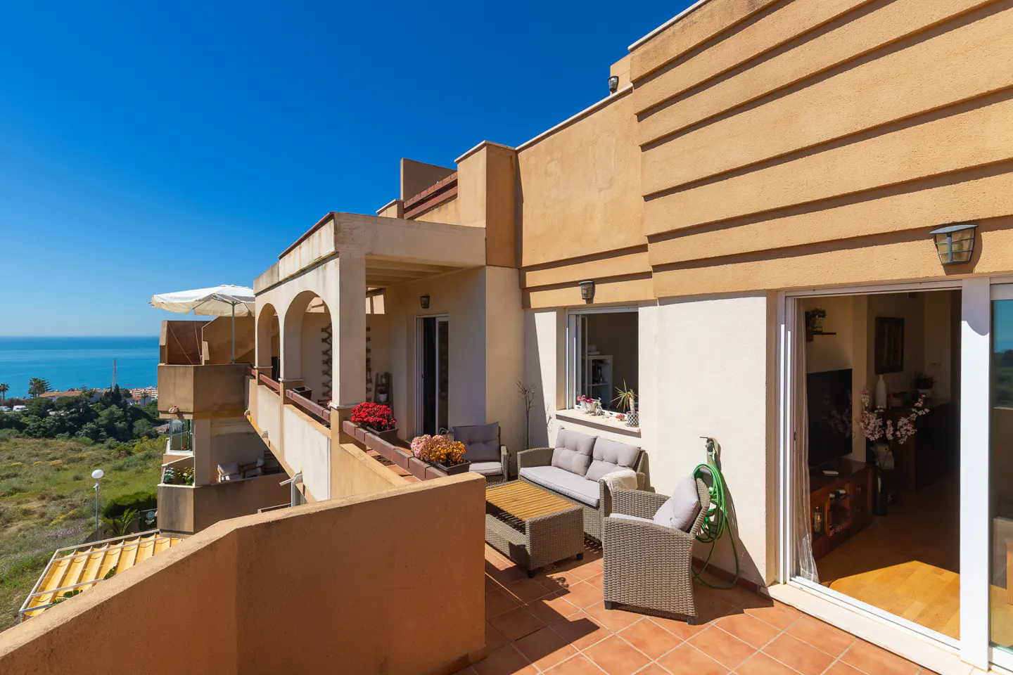Balcony with outdoor seating and ocean view. Beige building with arched openings and a tiled terrace. Blue sky and green landscape in the background.