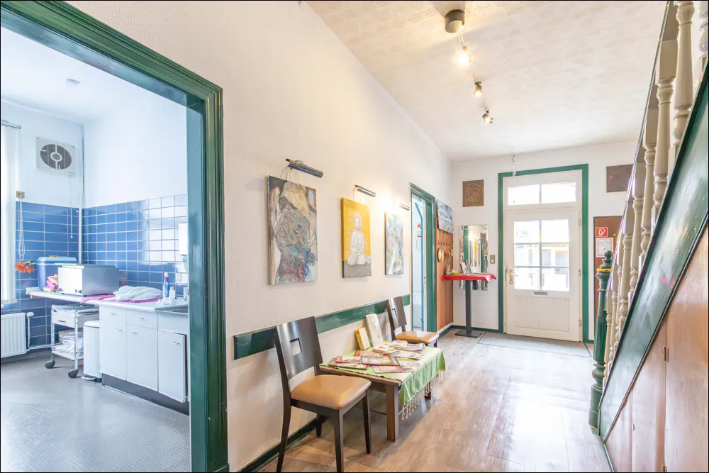 Hallway with white walls, green trim, and wood floors. Art hangs on the wall above a table and chairs. A kitchen is visible through a doorway.