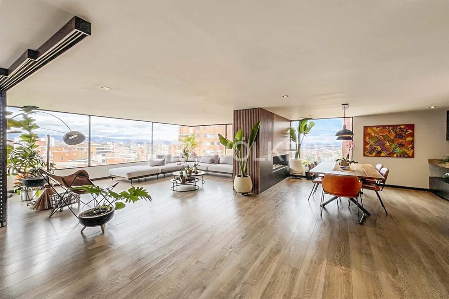 Bright, open-concept living space with wood floors, floor-to-ceiling windows, and city views. Features a gray sofa, dining table, and indoor plants.