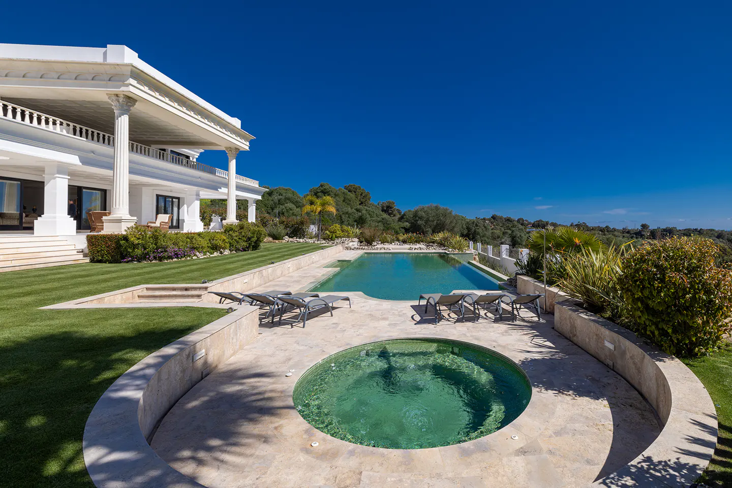 Luxury home exterior with a pool, hot tub, and lounge chairs on a sunny day. The house is white with columns.