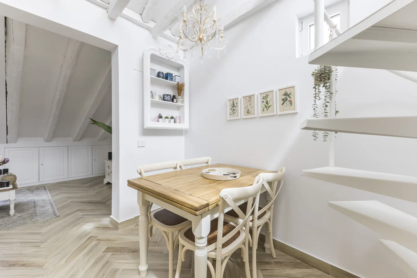 Bright, all-white dining area with a wooden table, four chairs, and a chandelier. A spiral staircase and botanical prints add charm.