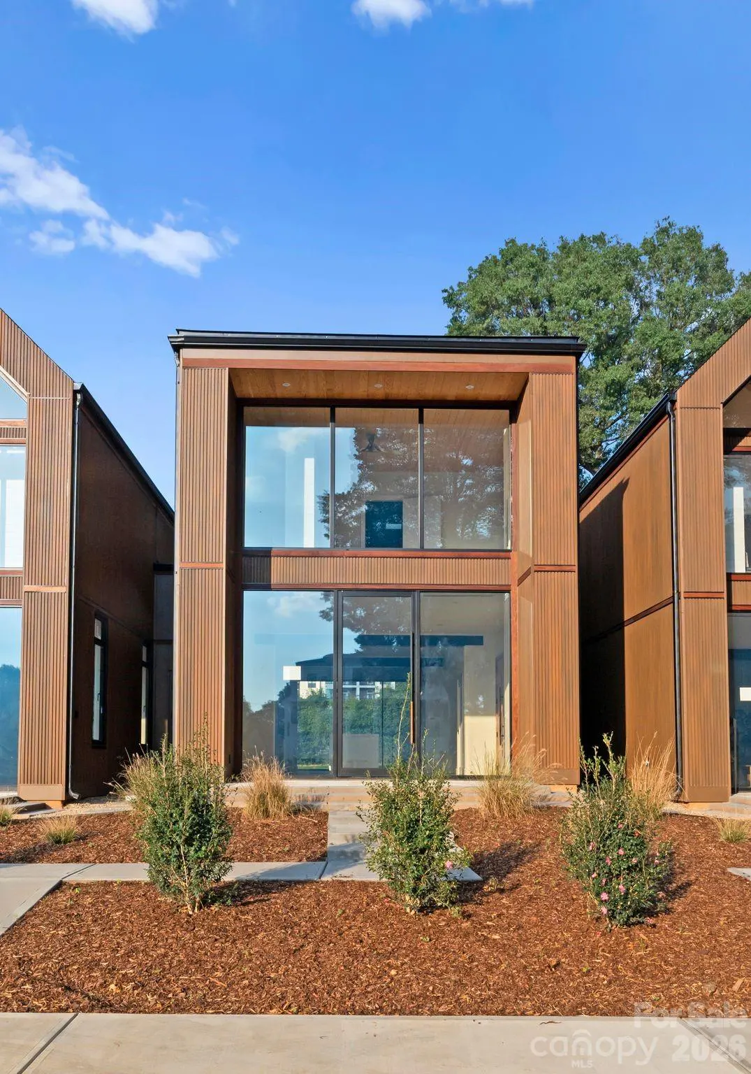 Modern two-story home with vertical brown siding, large glass windows, and a landscaped front yard with mulch.