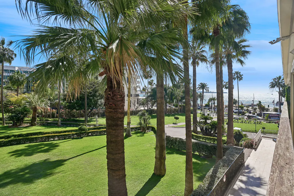 View from a balcony of palm trees, green lawn, and the ocean under a blue sky.