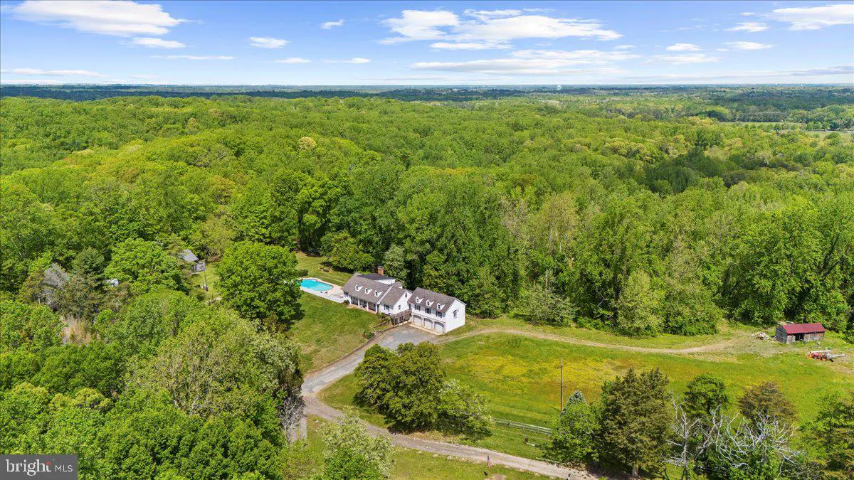 Aerial view of a white house with a pool, surrounded by lush green trees under a blue sky with scattered clouds.