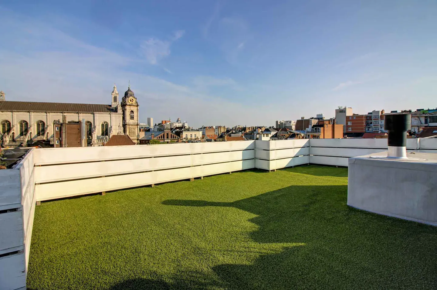 Rooftop terrace with artificial grass, white fence, and city views under a blue sky.