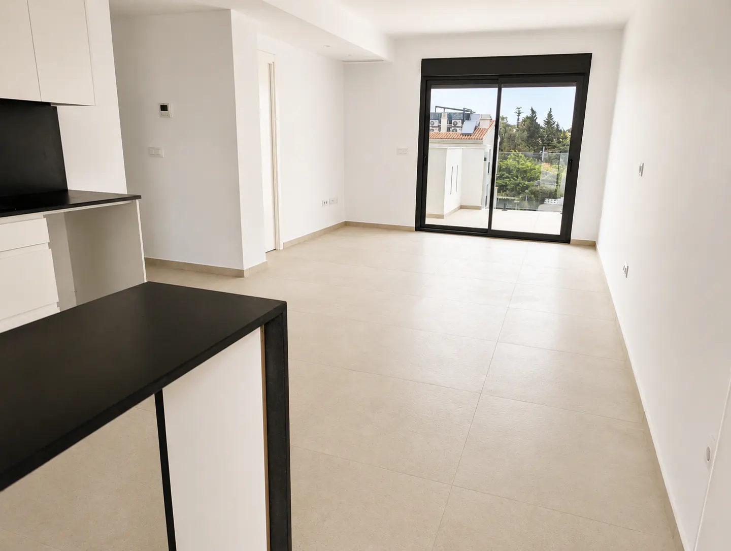 Bright, empty room with beige tile floor, white walls, and black-framed sliding glass doors to a balcony. Kitchen counter in foreground.