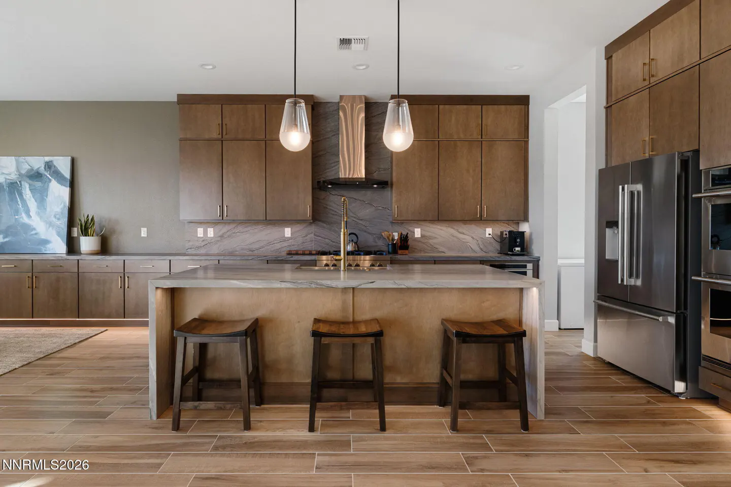 A modern kitchen with wood cabinets, a marble countertop island with three stools, and stainless steel appliances.