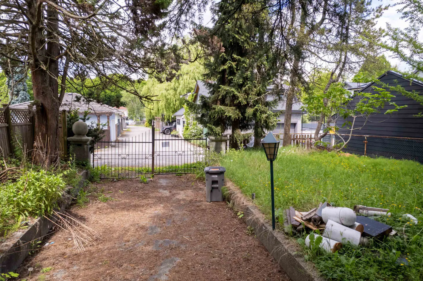 View of a property's gated entrance, with a lamp post, trash bin, and debris on the overgrown lawn.